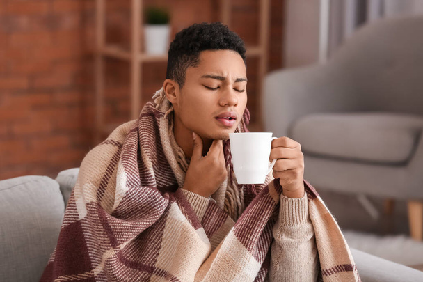 Ill African-American guy with sore throat drinking hot tea at home - 写真・画像