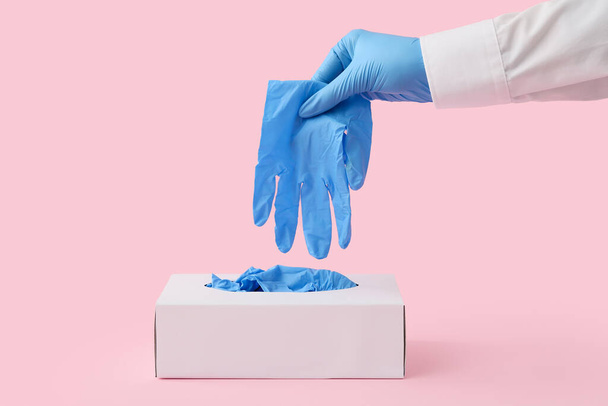 Γιατρός λαμβάνοντας μπλε ιατρικό γάντι από το κουτί σε ροζ φόντο - Φωτογραφία, εικόνα