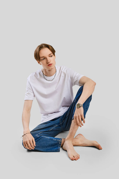 Young barefoot man sits on the floor in studio in t-shirt and jean - Valokuva, kuva