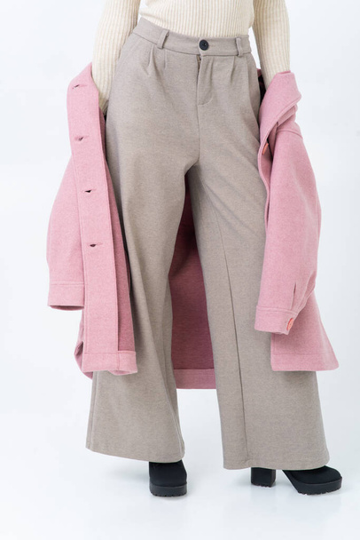 The woman takes off her pink coat, wearing a beige warm turtleneck, gray wide trousers and black heeled shoes. Without a face. White background. - Foto, afbeelding