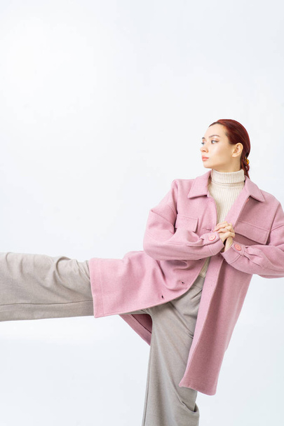 A red-haired Asian woman poses in a pink coat, beige turtleneck, gray pants. The leg is raised up, the hands are folded together. Warm fashionable autumn clothes. White background. Looks away. - Photo, Image