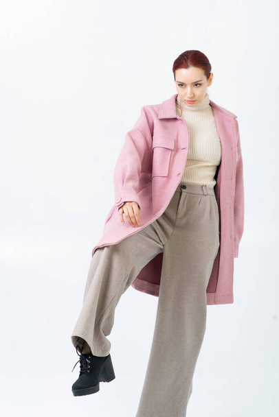 A red-haired Asian woman poses in the studio in a pink coat, beige turtleneck, gray pants and black shoes. Warm fashionable autumn clothes. White background. Looks away. - Foto, afbeelding