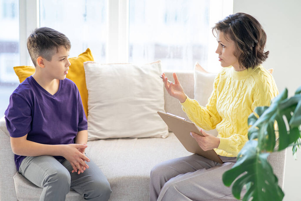 Teenager boy is undergoing therapy with middle-aged woman psychologist. They sit in cozy interior on sofa near window. The horizontal position of frame. Blurred foreground. - Foto, Imagen