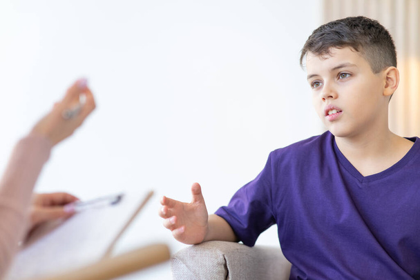 Teenage boy in purple T-shirt is gesturing and sharing his experiences with psychotherapist. Female hand with pen in foreground blurred. - Valokuva, kuva