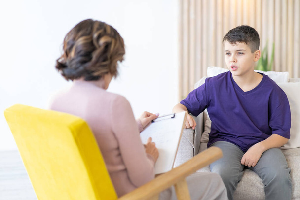 Teenage boy in purple T-shirt comparte sus experiencias con el psicoterapeuta. Mujer psicoterapeuta en silla amarilla en primer plano borrosa. - Foto, imagen