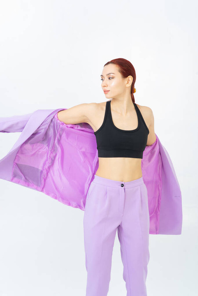 A woman puts on a purple suit jacket with a sporty black top. Red hair. Light background. Posing in the studio. - Photo, Image