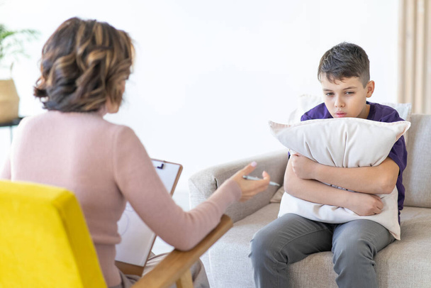 Worried teenager in purple T-shirt hugs pillow while sitting on couch with psychologist for consultation. Psychologist in chair in front of blurred background. - 写真・画像