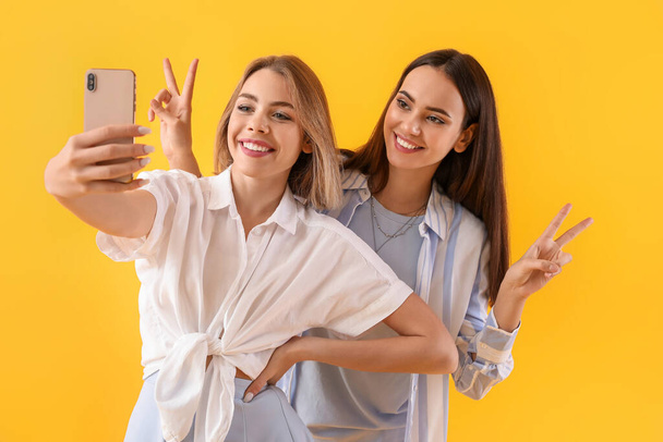 Young women with mobile phone taking selfie on yellow background - Foto, Imagen