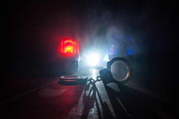 Полицейский рейд ночью, и вы арестованы. Силуэт наручников с полицейской машиной сзади. Изображение с мигающими красными и синими полицейскими огнями на туманном фоне. Селективный фокус - Фото, изображение