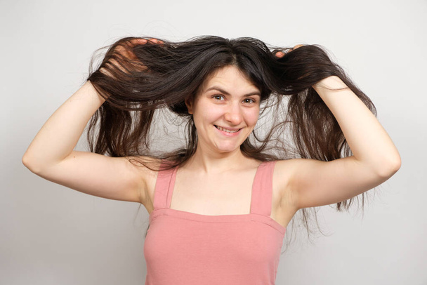 Krásná brunetka žena s dlouhými vlasy na bílém pozadí drží ruce na hlavě a přemýšlí o tom, co účes dělat - Fotografie, Obrázek