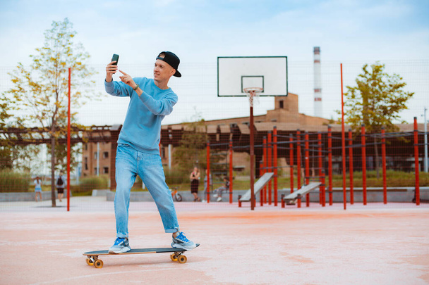 Full-leghth of young hipster guy in cap makes selfie with mobile phone while sitting on skateboard, gestures victory sign with fingers, skateboarder sharing photos with followers on social networks - Foto, imagen
