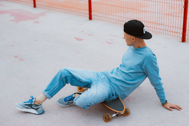 Portrait of young hipster man in stylish cap standing with wooden skateboard on shoulder, looking away thoughtfully, spending time actively outdoors in city park. Skateboarding sport concept - Photo, Image