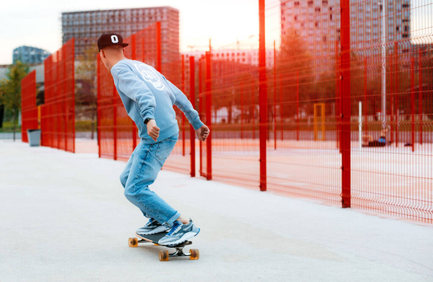 Rear view of teenager riding skateboard along red high fence at skatepark, millennial guy skating on longboard spending time actively outdoors on summer evening. Extreme hobby and skateboarding - Photo, Image