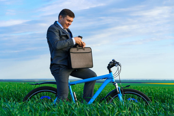 ビジネスコンセプト-ビジネスマンは緑の芝生のフィールドに自転車でポーズ、ビジネススーツを着て、彼はブリーフケースの中に何かを探しています - 写真・画像
