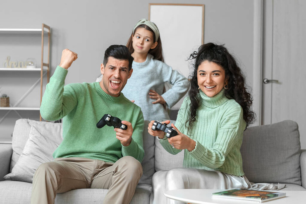 Маленька дочка і її батьки в теплих светрах грають у відеоігри вдома
 - Фото, зображення