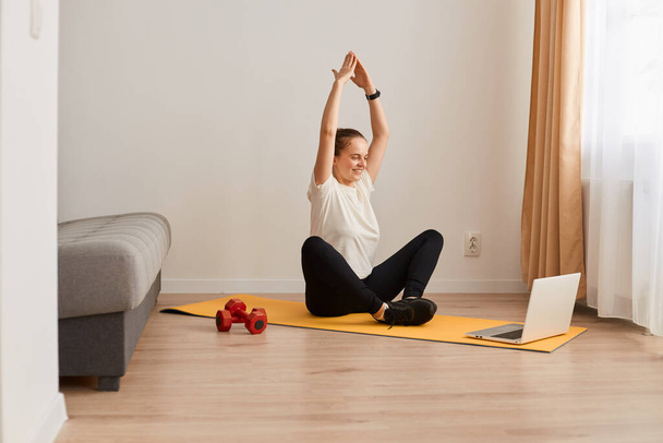 Portrait of sporty girl sitting on mat and using laptop for online workout, wearing white T-shirt and black leggins , doing yoga via online lesson, posing with raised arms. - Φωτογραφία, εικόνα