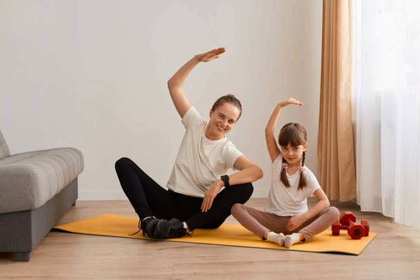 Indoor shot of slim woman with hair bun sitting in lotus position on mat practicing yoga with her cute female child, bending to side with raised hand, doing stretching workouts with daughter. - Zdjęcie, obraz