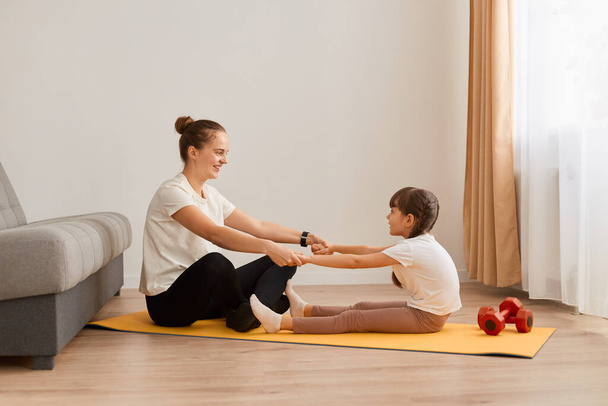 Indoor shot of woman mother doing sport with her little daughter, sitting on yoga mat on floor and holding hands, looking at each other, fitness together at home. - Foto, imagen