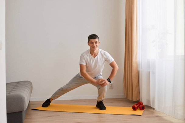 Indoor shot of happy athletic man wearing sportswear doing lower body sport exercise, stretching legs, warming up training muscles for flexibility, looking at camera with smile. - Foto, Imagen