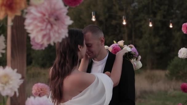 A charming portrait of the newlyweds who embrace and the groom kisses the bride at the arch with dahlias. Slow motion - Footage, Video