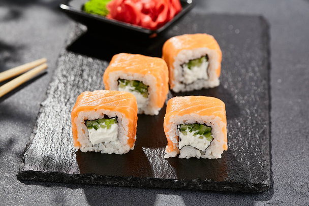 Maki sushi on dark stone table. Philadelphia maki with salmon. Sushi roll with cheese, cucumber inside, salmon outside. Style concept japanese menu with black background, leaves and hard shadow - Valokuva, kuva