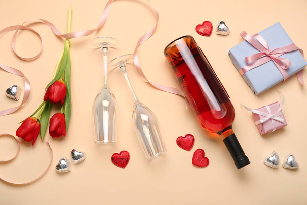 Σύνθεση με νόστιμες καραμέλες σε σχήμα καρδιάς, λουλούδια και μπουκάλι κρασί για τον εορτασμό της ημέρας του Αγίου Βαλεντίνου σε μπεζ φόντο - Φωτογραφία, εικόνα
