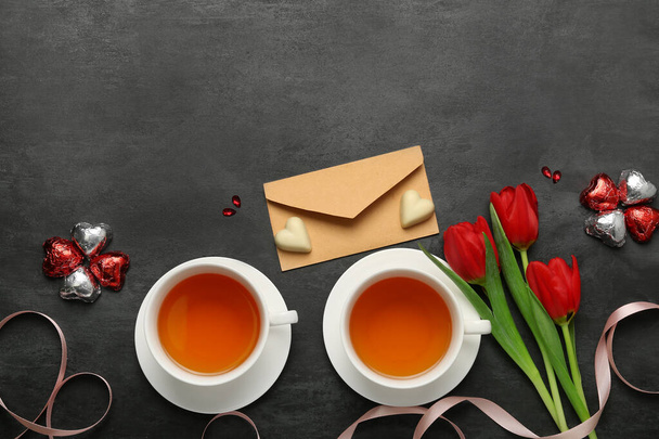 Σύνθεση με νόστιμες καραμέλες σε σχήμα καρδιάς, φάκελο, φλυτζάνια τσάι και λουλούδια για τον εορτασμό της ημέρας του Αγίου Βαλεντίνου σε σκούρο φόντο - Φωτογραφία, εικόνα