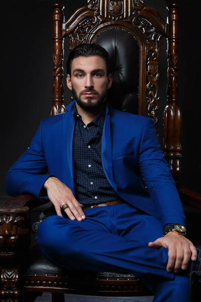 Elegant man in a classic blue suit sitting in a carved wooden chair against a dark background. - Fotó, kép