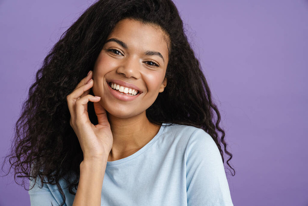Fiatal fekete nő göndör haj mosolyog, és néz kamera elszigetelt felett lila háttér - Fotó, kép