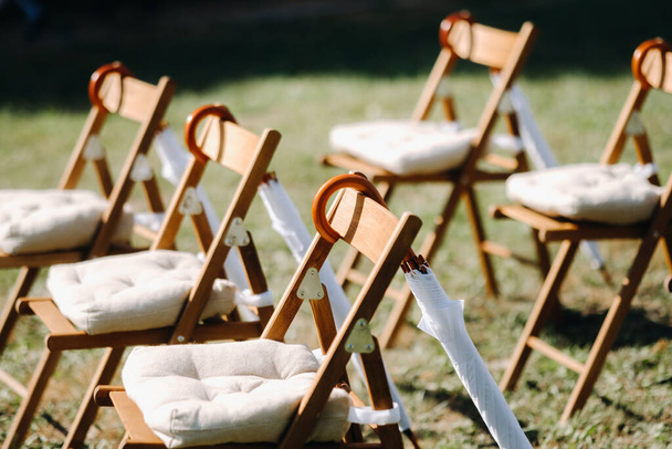 Chairs stand on the green grass in the area of the wedding ceremony, white umbrellas hang on the chairs. - Photo, Image