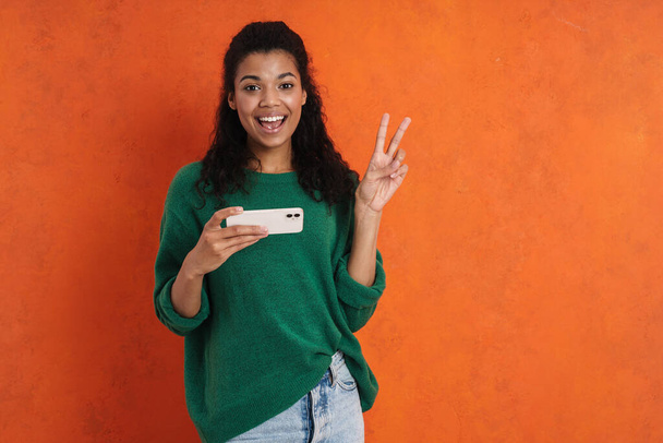 Happy smiling african casual brunette woman standing over orange wall background, holding mobile phone, playing games, watching video - Foto, Bild