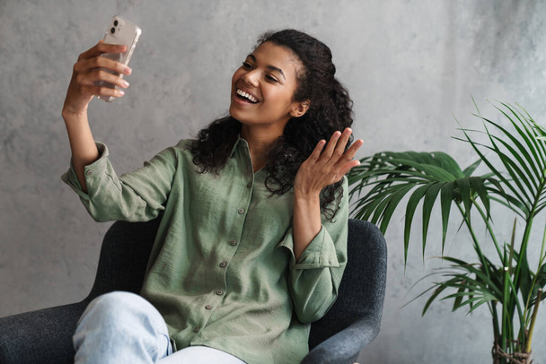 Black young woman taking selfie on mobile phone while sitting in armchair indoors - Foto, imagen