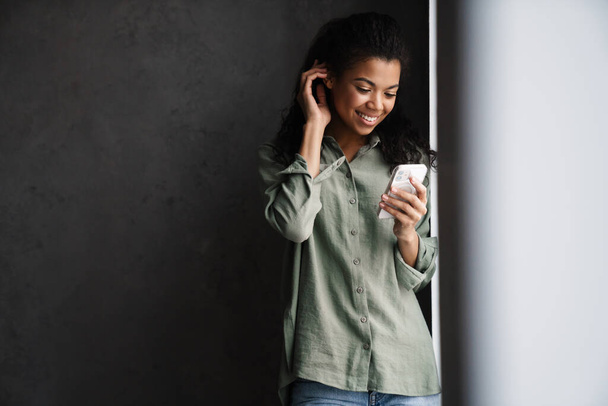 Black young woman using mobile phone while leaning on wall indoors - Photo, image