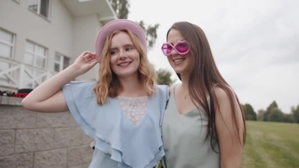 Jedna młoda dziewczyna w kapeluszu i druga w śmiesznych okularach zabawia się przed kamerą i grymasem pokazując język  - Materiał filmowy, wideo