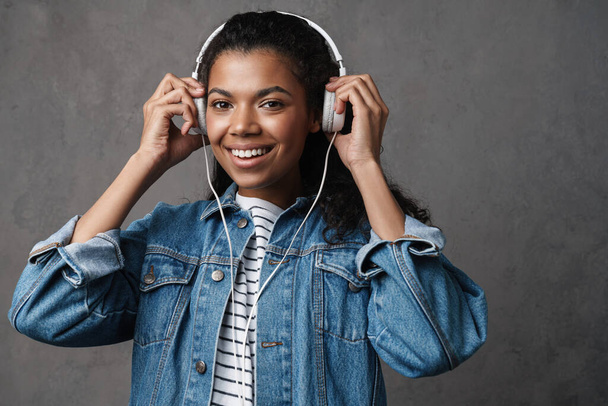 Happy young african woman in casual wear standing over gray background, listening to music with headphones - Valokuva, kuva