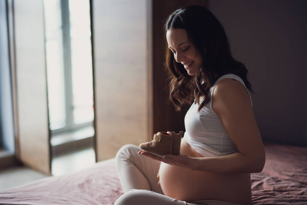 Pregnant woman relaxing at home. She is sitting on bed and holding baby shoes. - 写真・画像