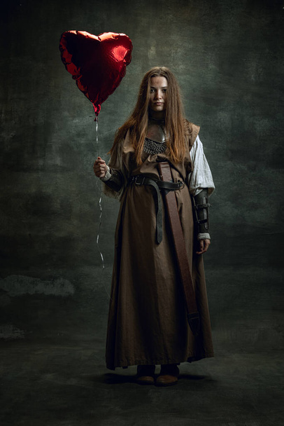 Full-length portrait of sad beautiful woman in image of medieval warrior or knight with dirty wounded face with heart shape balloon isolated over dark vintage background. - Fotografie, Obrázek