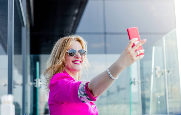 A successful woman entrepreneur in a magenta jacket makes a selfie on the terrace of a cafe in a business center. Business woman representation concept - Photo, Image