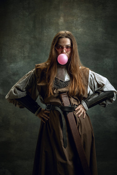 Portrait of adorable woman, medieval female warrior or knight with dirty wounded face looking at camera isolated over dark retro background. Funny meme emotions - Фото, изображение