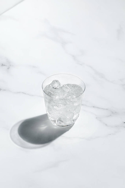 Glass of cold water with ice on sunny marble background, dark shadow. Creative concept - Фото, изображение