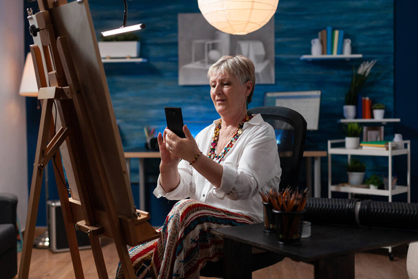 Пожилой художник, сидящий перед мольбертом с холстом, просматривающим смартфон - Фото, изображение