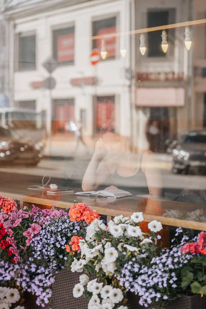 Behind window glass. Girl at table with book, reflection of city street on summer day, blurred bokeh - Φωτογραφία, εικόνα