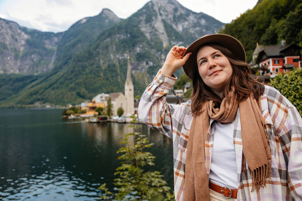 Hallstatt, Avusturya: Güzel romantik kadın turistin portresi, doğal ışık, vücut pozitifliği, bej renkli beyaz çizgili kız, şapka ve fular, manzara resimli şehir sokağı - Fotoğraf, Görsel