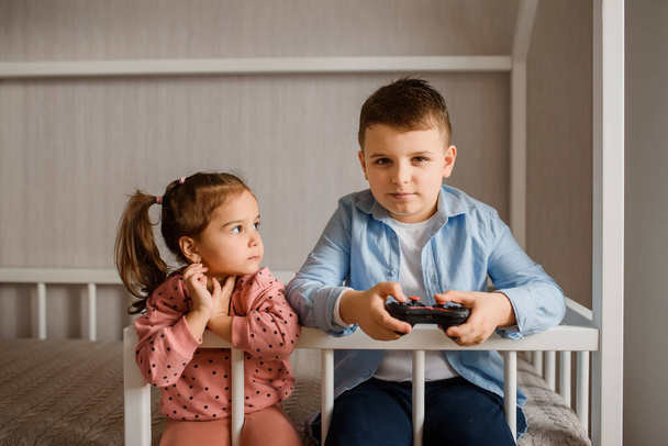 Two Children brother and sister are sitting in a Montessori bed and playing Video games. Boy is holding a gamepad and girl is looking to him. - Fotoğraf, Görsel