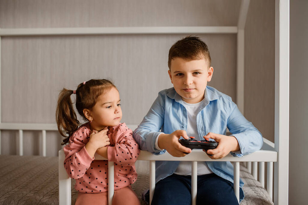 Two Children brother and sister are sitting in a Montessori bed and playing Video games. Boy is holding a gamepad and girl is looking to him. - Valokuva, kuva