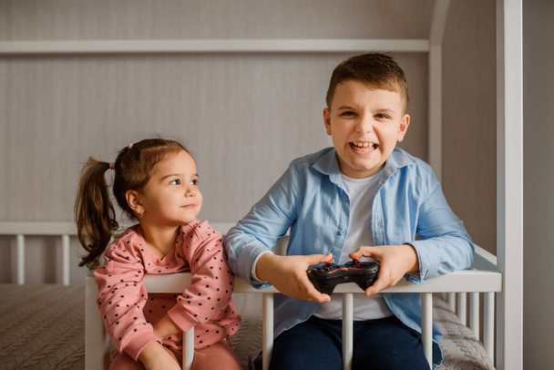Dos niños hermano y hermana están sentados en una cama Montessori y jugando Videojuegos. Boy está sosteniendo un mando y la chica lo está mirando. - Foto, Imagen