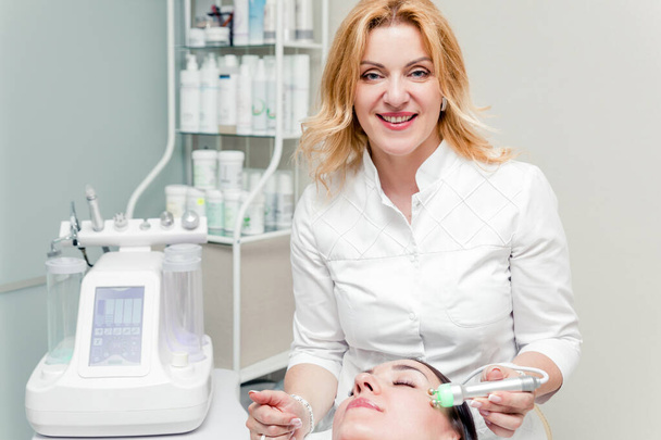 Косметолог робить пацієнту процедуру мікрострумової терапії. Поруч з косметологом знаходиться косметична машина
 - Фото, зображення