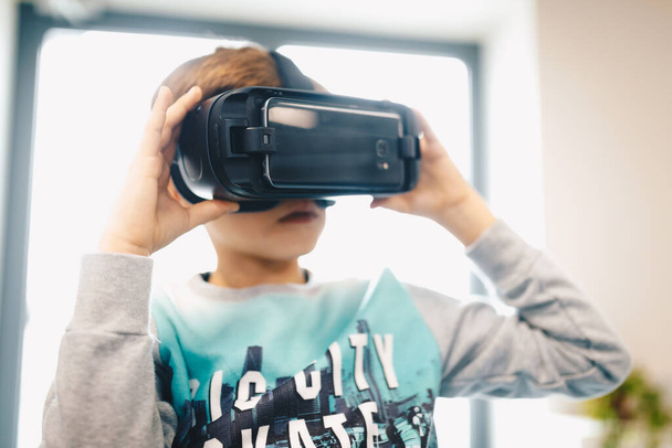 Kinder erleben virtuelle Realität in der Schule. Erstauntes Kind mit Virtual-Reality-Headset, das zu Hause hinter einem Tisch sitzt. Überraschter kleiner Junge schaut in VR-Brille - Foto, Bild