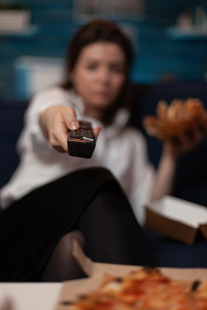 Vista de cerca del control remoto en la mano de la mujer cambiando los canales de televisión mientras come comida rápida para llevar hamburguesa - Foto, Imagen