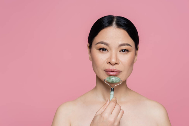 Mulher asiática com ombros nus massageando queixo com rolo de jade isolado em rosa  - Foto, Imagem